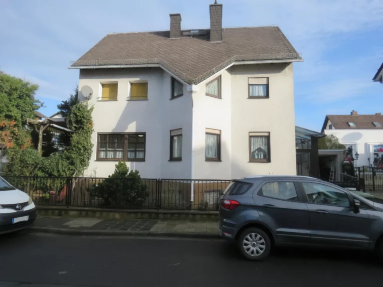 Ansicht eines zweistöckigen Einfamilienhauses mit Garten und Garage an einer Straße.