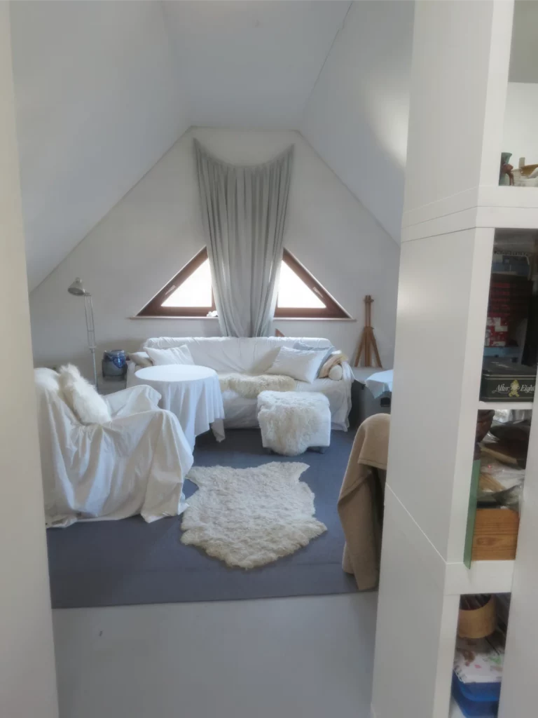 Helles Dachzimmer mit weißen Möbeln, Teppich, Sofa und Fenster mit Vorhängen für ein gemütliches Ambiente.